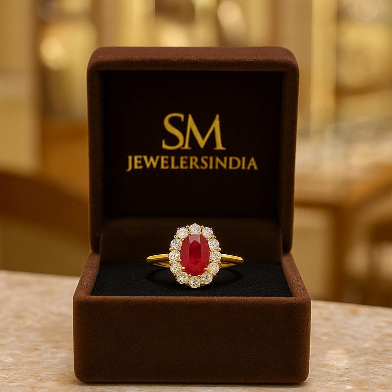 Op de afbeelding: Een gouden ring met een grote ovale robijn als middensteen, omringd door een halo van kleinere diamanten. De ring wordt gepresenteerd in een bruine sieradendoos met de tekst "SM JEWELERSINDIA" in gouden letters.