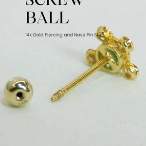 May include: A gold screw ball nose piercing with a small green stone in the center. The screw ball is surrounded by small clear stones. The piercing is 14k gold.
