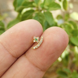 May include: A gold nose ring featuring a crescent moon design with a small green gemstone at the centre of a four-pointed star.