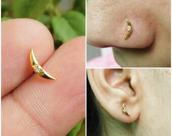 May include: A gold-colored crescent moon earring with a small, clear gemstone. The earring is shown in multiple views, including on a finger, in a nose piercing, and in an ear piercing. The jewelry is small and delicate.