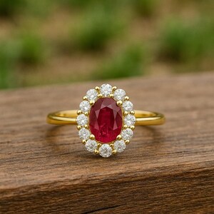Op de afbeelding: Een gouden ring met een grote ovale robijn als middensteen, omringd door een halo van kleinere, ronde diamanten. De ring is gezet op een dunne, gepolijste gouden band, die het levendige rood van de robijn en de schittering van de diamanten benadrukt. De ring ligt op een houten oppervlak.