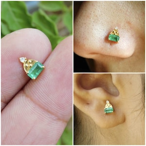 May include: A pair of gold earrings featuring a small emerald green gemstone and a single diamond accent. The earrings are pictured on a person's ear and on a finger.