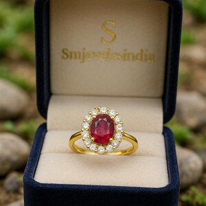 Op de afbeelding: Een gouden ring met een grote ovale robijn in het midden, omringd door een halo van kleine diamanten. De ring wordt gepresenteerd in een donkerblauwe fluwelen ringdoos. De tekst "Smjevelsindia" is zichtbaar.