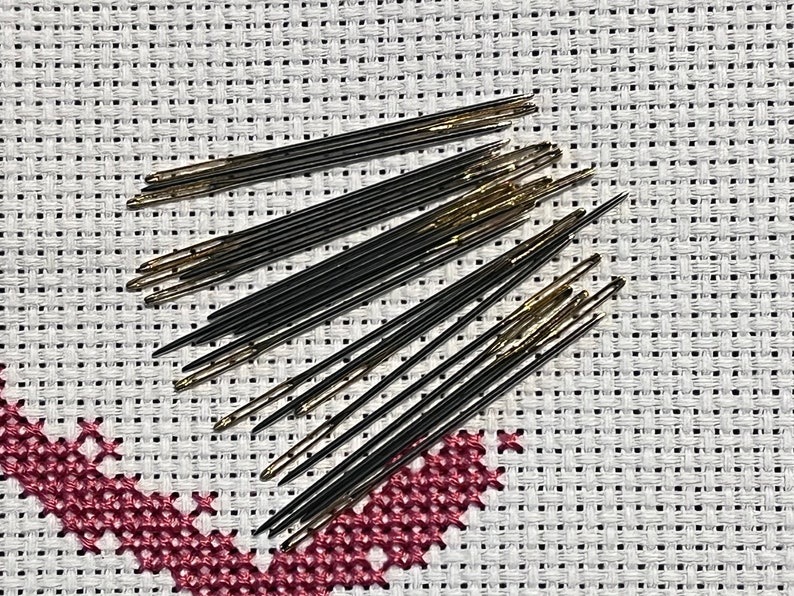 May include: A close-up of a group of black and gold embroidery needles on a white Aida cloth. The needles are arranged in a fan shape.