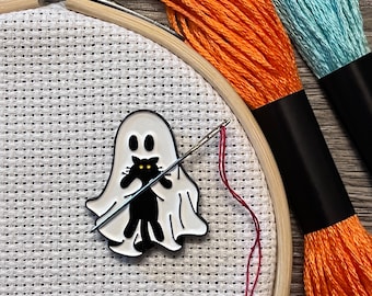 May include: A white ghost with a black cat inside is being stitched on white Aida cloth in a wooden embroidery hoop. Orange and blue embroidery floss is visible in the background.