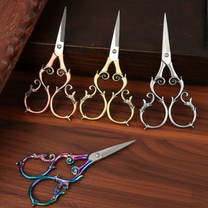 May include: Four decorative scissors with ornate handles and pointed blades. The scissors come in rose gold, gold, silver, and a rainbow iridescent finish. The handles feature a scroll and leaf design, and the blades are silver.