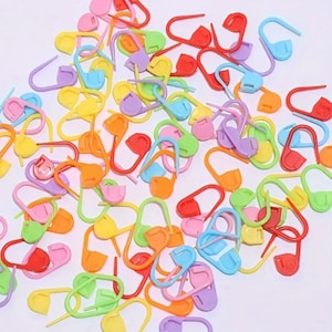 May include: A large assortment of colorful plastic stitch markers. The markers are shaped like a small loop with a pointed end. The colors include red, orange, yellow, green, blue, purple, and pink.