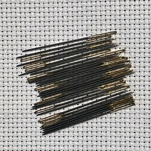 May include: A close-up of a bundle of black and gold embroidery needles on a white Aida cloth.