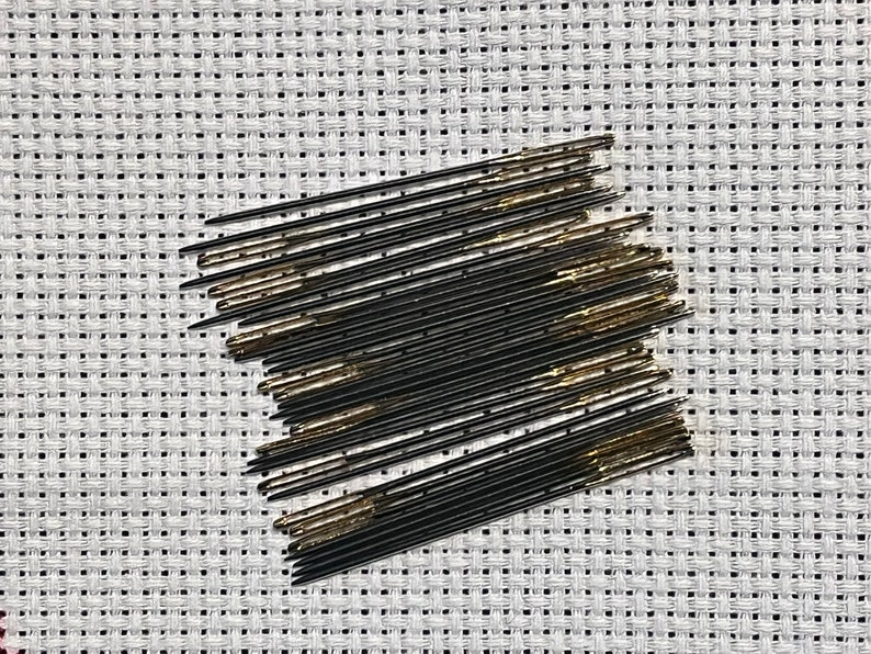 May include: A close-up of a bundle of black and gold embroidery needles on a white Aida cloth.