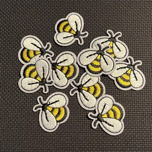 May include: A collection of embroidered bee patches. Each patch features a white outline, yellow and black striped body, and white wings. The patches are scattered on a dark gray textured surface.