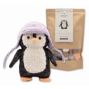 May include: A handmade knitted penguin wearing a lavender hat with ear flaps. The penguin has a white belly, black body, and orange feet and beak. A craft kit with yarn and instructions is in the background. The kit's label reads "Jack Penguin".