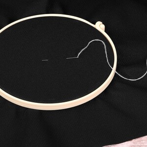 May include: A cream-colored embroidery hoop holds taut black fabric, with a needle and white thread in the center. The needle is threaded, and a length of thread extends from the fabric, suggesting the start of a project.