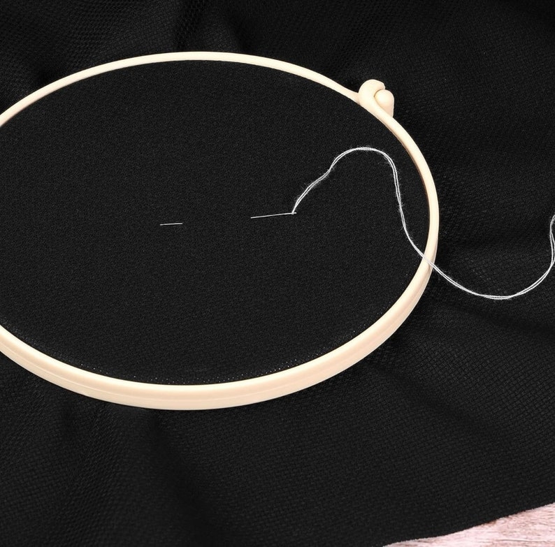 May include: A cream-colored embroidery hoop holds taut black fabric, with a needle and white thread in the center. The needle is threaded, and a length of thread extends from the fabric, suggesting the start of a project.
