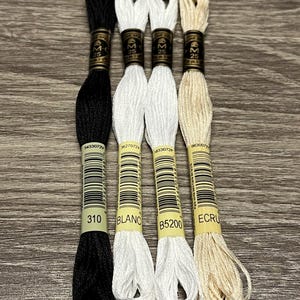 May include: Four spools of embroidery floss in black, white, white, and beige. The spools are labeled with the DMC numbers 310, BLANC, B5200, and ECRU.