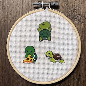 May include: Three enamel pins featuring cartoon turtles. One turtle is green and has a shell with a green and black pattern. Another turtle is green and is holding a slice of pizza. The third turtle is green and brown and is holding a piece of seaweed. The pins are displayed on a white fabric background.