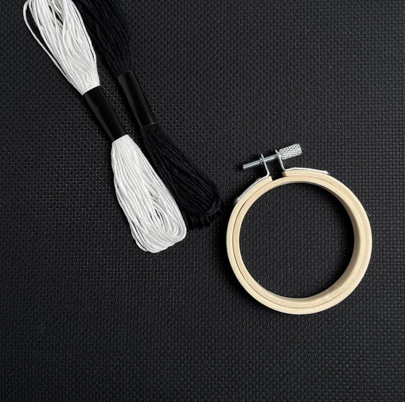 May include: A craft image featuring a wooden embroidery hoop, white and black embroidery floss. The hoop is round with a metal tightening mechanism. The floss is neatly bundled. The background is a textured black surface.