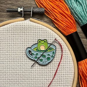 May include: A green frog sitting in a blue teacup with green leaves on the side. The teacup is on a white Aida cloth stretched in a wooden embroidery hoop. A red thread is threaded through a needle and is partially visible in the hoop.