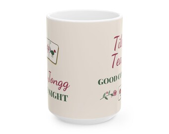 Può includere: Una tazza in ceramica color crema con illustrazioni di tessere del Mahjong, testo rosso e verde che recita Tile Tea, Jongg Night e GOOD COMPANY, insieme a una piccola grafica floreale e una singola tessera del Mahjong.