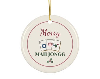 May include: A round, cream-colored ceramic ornament with a gold hanging string. It features the text Merry MAH JONGG, an illustration of three mahjong tiles, and a small holly sprig. A thin maroon circular border frames the design.