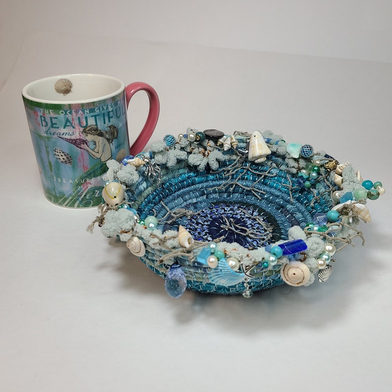 K&ouml;nnte beinhalten: Eine blaue und wei&szlig;e gewebte Schale mit verschiedenen maritimen Verzierungen, darunter Muscheln, Perlen und eine kleine wei&szlig;e Figur. Die Schale steht auf einer wei&szlig;en Oberfl&auml;che. Der Text auf der Tasse lautet "The ocean gives beautiful dreams treasure"