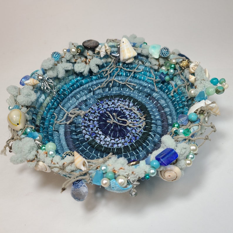 K&ouml;nnte beinhalten: Eine blaue und wei&szlig;e gewebte Schale mit einer Vielzahl von Meeresmotiven, darunter Muscheln, Perlen und ein kleines wei&szlig;es Keramikornament. Die Schale ist mit einem blauen und wei&szlig;en Wirbelmuster verziert.