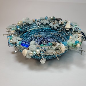 K&ouml;nnte beinhalten: Eine blaue und wei&szlig;e gewebte Schale, verziert mit Muscheln, Perlen und anderen maritimen Verzierungen.