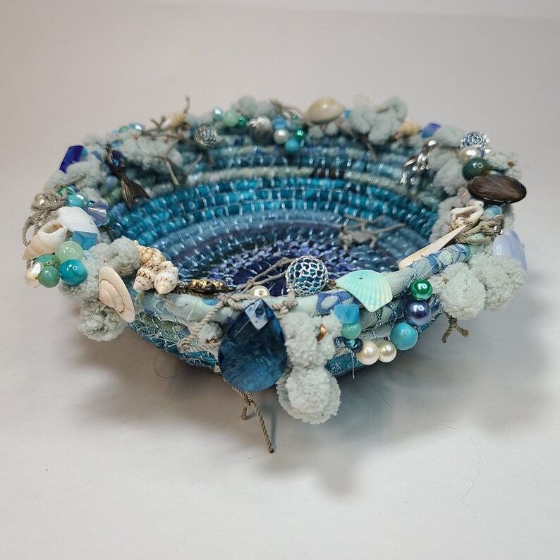 K&ouml;nnte beinhalten: Eine blaue und wei&szlig;e gewebte Schale, verziert mit Muscheln, Perlen und Bommeln. Die Schale hat einen blauen und wei&szlig;en Ombre-Effekt.