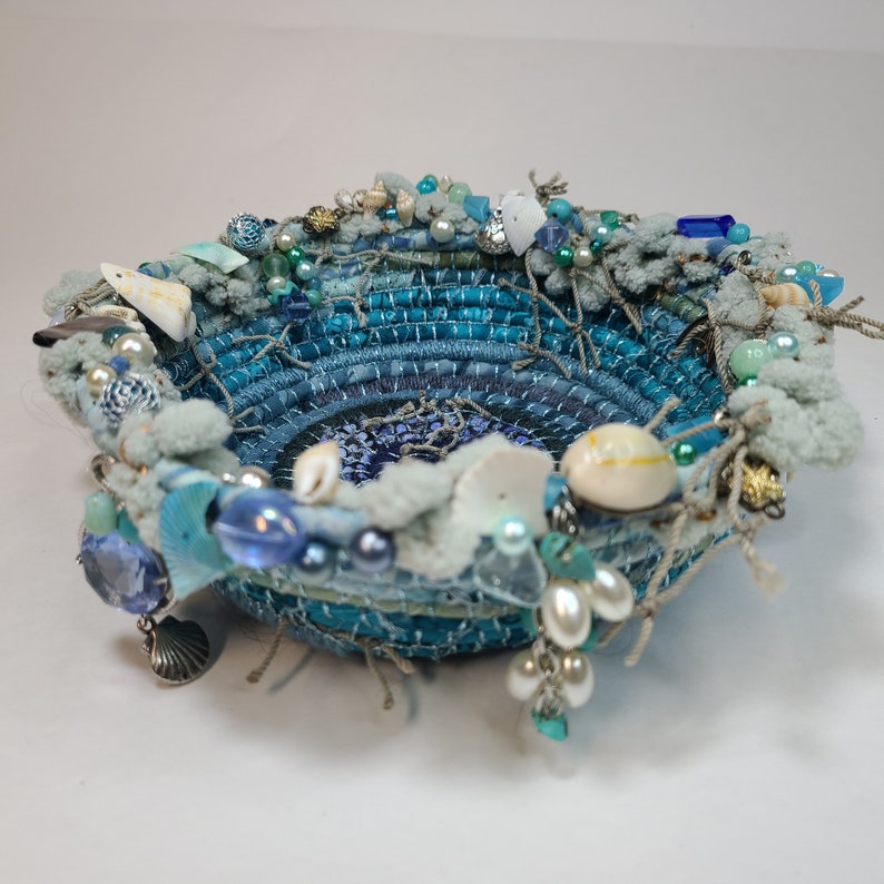 K&ouml;nnte beinhalten: Eine blaue und wei&szlig;e gewebte Schale, verziert mit Muscheln, Perlen und anderen maritimen Verzierungen.