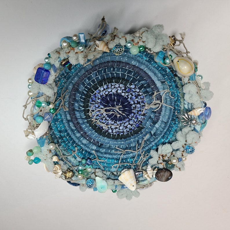 K&ouml;nnte beinhalten: Ein blauer und wei&szlig;er runder Wandbehang mit einem gewebten Zentrum und verschiedenen Verzierungen, darunter Muscheln, Perlen und Stoffblumen.