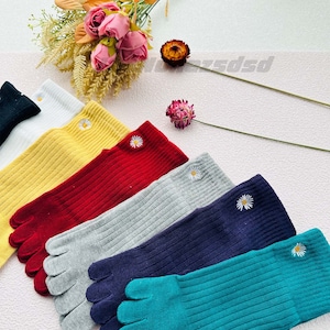 May include: Six pairs of colourful toe socks with a white daisy embroidered on each sock. The socks are arranged in a row from left to right, with the colours being black, white, yellow, red, grey, navy blue, and teal.