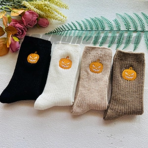 May include: Four pairs of socks in black, white, beige, and brown, each with an embroidered orange jack-o'-lantern design. The socks are made of a ribbed knit material, ideal for autumn and Halloween.