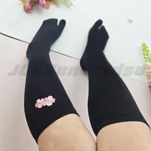 Puede incluir: Un par de calcetines negros con punta dividida que llegan por encima de la rodilla. Los calcetines cuentan con un compartimento separado para el dedo gordo y están decorados con un pequeño diseño floral bordado en rosa y blanco en el lado exterior.