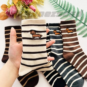 Puede incluir: Conjunto de cuatro pares de calcetines a rayas con un dachshund bordado en cada calcetín. Los calcetines son de color marrón, negro y blanco.