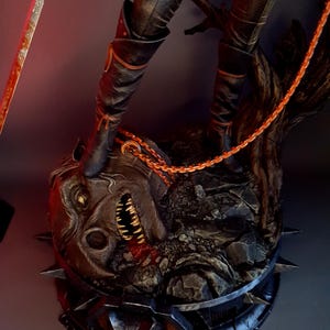 May include: A detailed statue featuring a figure in black boots standing on a grotesque, monster-like head. The head is dark with sharp teeth and spikes. An orange chain and a sword are also visible, adding to the dark fantasy theme.