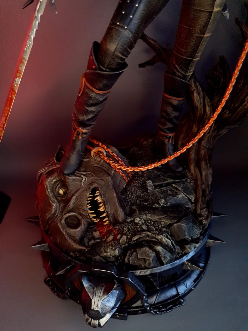 May include: A detailed statue featuring a figure in black boots standing on a grotesque, monster-like head. The head is dark with sharp teeth and spikes. An orange chain and a sword are also visible, adding to the dark fantasy theme.