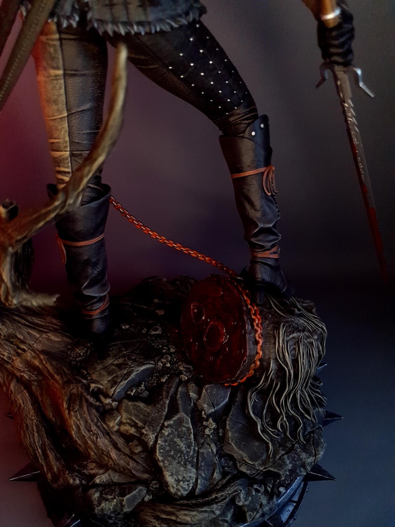 May include: A detailed figurine of a character in black leather pants and boots, adorned with white dots. The figure holds a sword and stands on a textured, rocky base. A red circular object with a chain is also present.