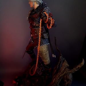 May include: A detailed figurine of a character from The Witcher series. The figure is clad in black and red armor, holding an orange braided whip. The character has silver hair and is posed on a detailed, textured base.