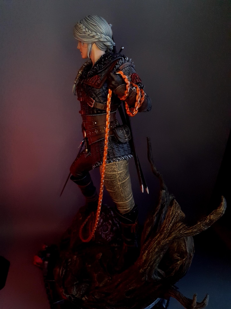 May include: A detailed figurine of a character from The Witcher series. The figure is clad in black and red armor, holding an orange braided whip. The character has silver hair and is posed on a detailed, textured base.