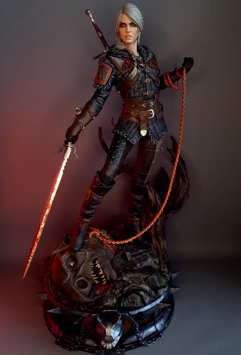 May include: A detailed statue of a woman in dark armor, holding a sword and a chain. The sword has a red hue, and the statue is standing on a monster's head. The statue is approximately 15 inches tall.