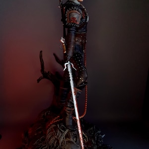 May include: A detailed statue of a character from The Witcher series. The figure is clad in dark armor and holds a silver sword with a blood-stained blade. The statue is mounted on a detailed base, with a sword strapped to the back.