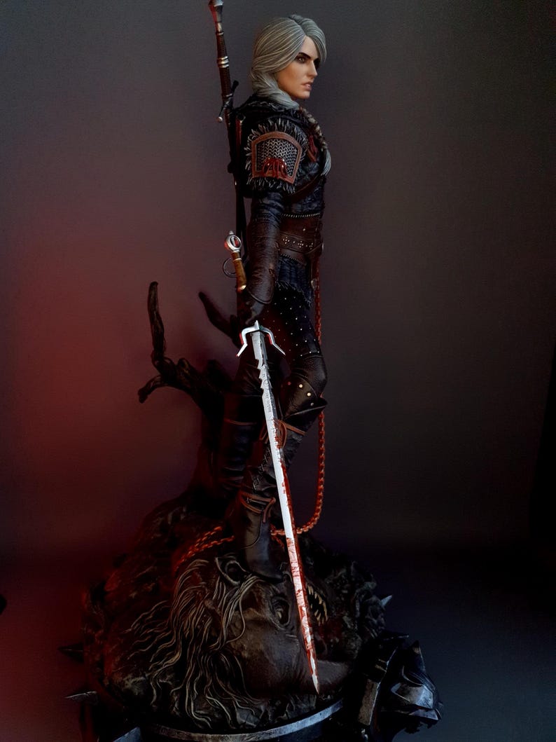 May include: A detailed statue of a character from The Witcher series. The figure is clad in dark armor and holds a silver sword with a blood-stained blade. The statue is mounted on a detailed base, with a sword strapped to the back.