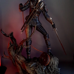 May include: A detailed figurine of a character from The Witcher series, clad in dark armor and wielding two swords. The figure stands on a detailed base with a tree and a severed head, with a chain attached to the character's belt.