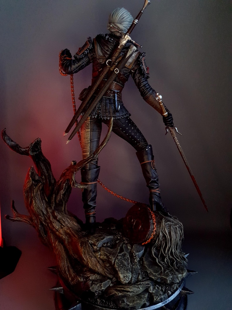 May include: A detailed figurine of a character from The Witcher series, clad in dark armor and wielding two swords. The figure stands on a detailed base with a tree and a severed head, with a chain attached to the character's belt.