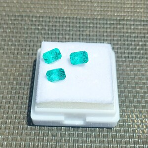 May include: Three faceted, rectangular, teal gemstones displayed on a white foam insert within a white plastic tray. The gemstones have a fan-like pattern carved into their surface. The background is a textured, gray surface.