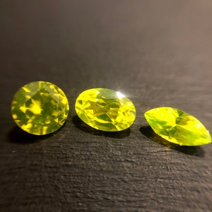 May include: Three green gemstones, cut and polished, arranged in a row on a dark surface.