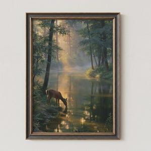 May include: A framed wall art print depicting a brown deer standing at the edge of a calm river in a dense forest. Sunlight filters through the tall green trees, creating a hazy atmosphere with reflections visible on the water surface.
