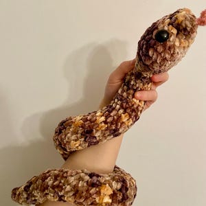 Op de afbeelding: Een pluche slangenknuffel in bruin-, paars- en geeltinten. De slang heeft een zwart knoopoog en een roze tong. Het zachte, getextureerde materiaal geeft het speelgoed een knuffelbaar uiterlijk. De slang is om een arm gewikkeld.