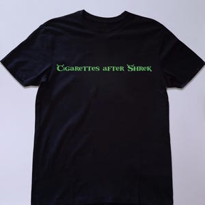 May include: Black t-shirt with the text "Cigarettes after Shrek" in green, stylized lettering. The shirt is a solid black color and appears to be made of a soft material. The text is centered on the chest.
