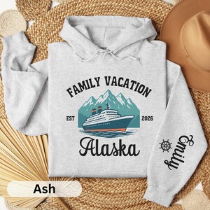 May include: Ash-colored hoodie with a "Family Vacation Alaska" design. The design includes a cruise ship, mountains, and the year 2026. The right sleeve features the name "Emily" and a ship's wheel.