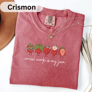 May include: A dusty rose-colored t-shirt featuring a graphic of five cartoon strawberries with various accessories and the white text "social work is my jam" printed below. The shirt has a crew neckline and a Comfort Colors brand label at the neck.